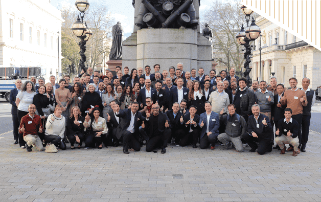 A large group of people gathered outside, all smiling and giving thumbs-up in front of a monument.